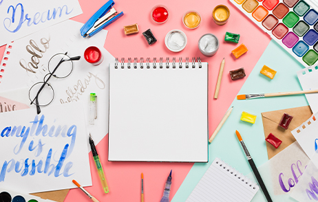 Bunte Stifte, ein Notizblock, Wasserfarben und Pinsel liegen auf einer bunten Unterlage