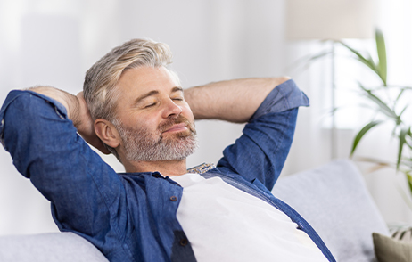 Ein Mann mittleren Alters mit grauen Haaren und Bart sitzt auf dem Sofa hat die Augen geschlossen und die Arme hinter dem Kopf verschränkt