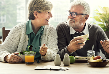 Ein älteres Paar sitzt zusammen beim Essen. Die beiden schauen sich fröhlich an.