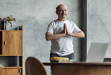 Älterer Mann mit Glatze steht hinter einem Schreibtisch und hat die Hände gefaltet. 