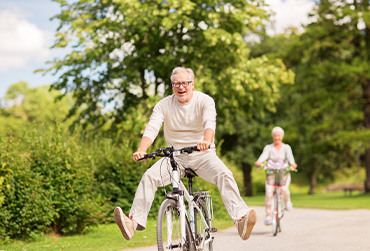 Älteres Paar fährt im Park Fahrrad. Der Mann fährt vorneweg und spreizt die Beine auf dem Fahrrad. Die Frau fährt hinterher.