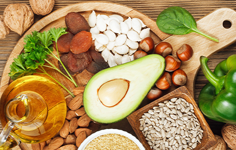 Verschiedene Nüsse, eine Avocado und eine Glaskaraffe mit Olivenöl liegen beziehungsweise stehen auf einem runden Holzbrett