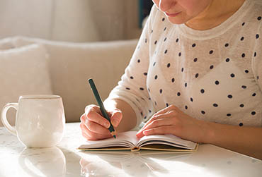 Eine Frau sitzt am Tisch und schreibt etwas in ihr Notizbuch. Neben ihr steht eine Tasse.