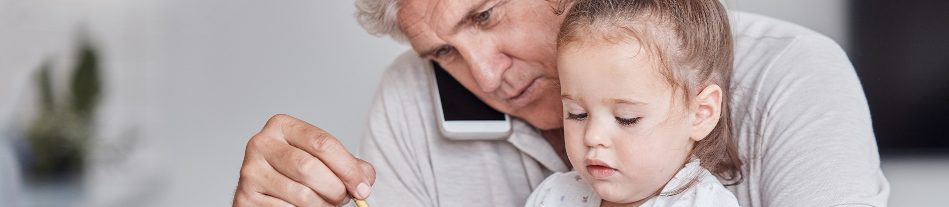 Der Opa hat die Enkelin auf dem Schoss sitzen. Er telefoniert mit dem Handy, hält einen Stift und hat den Laptop vor sich stehen.