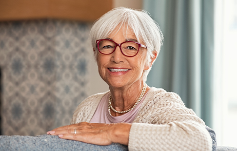 Eine ältere Frau mit weißen kurzen Haaren und roter Brille sitzt auf dem Sofa und lächelt freundlich