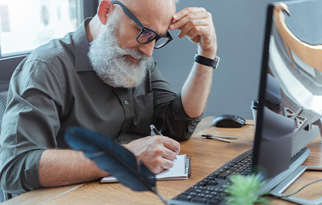 Ein älterer Mann mit weißem Bart und Brille sitzt am Schreibtisch und macht sich Notizen