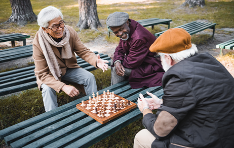 Drei ältere Männer sitzen im Park auf einer Bank. Zwei der Männer spielen Schach der andere schaut zu