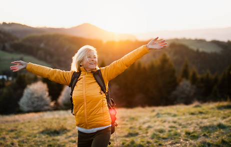 Eine Frau mittleren Alters mit gelber Jacke und Rucksack ist draußen in der Natur und streckt freudig die Arme nach oben