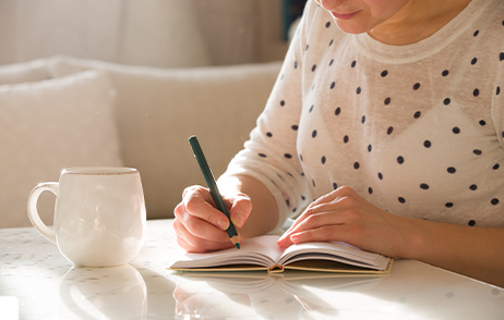 Eine Frau schreibt in ihr Tagebuch. Ihr Gesicht ist nicht erkennbar. Neben ihr steht eine Tasse.